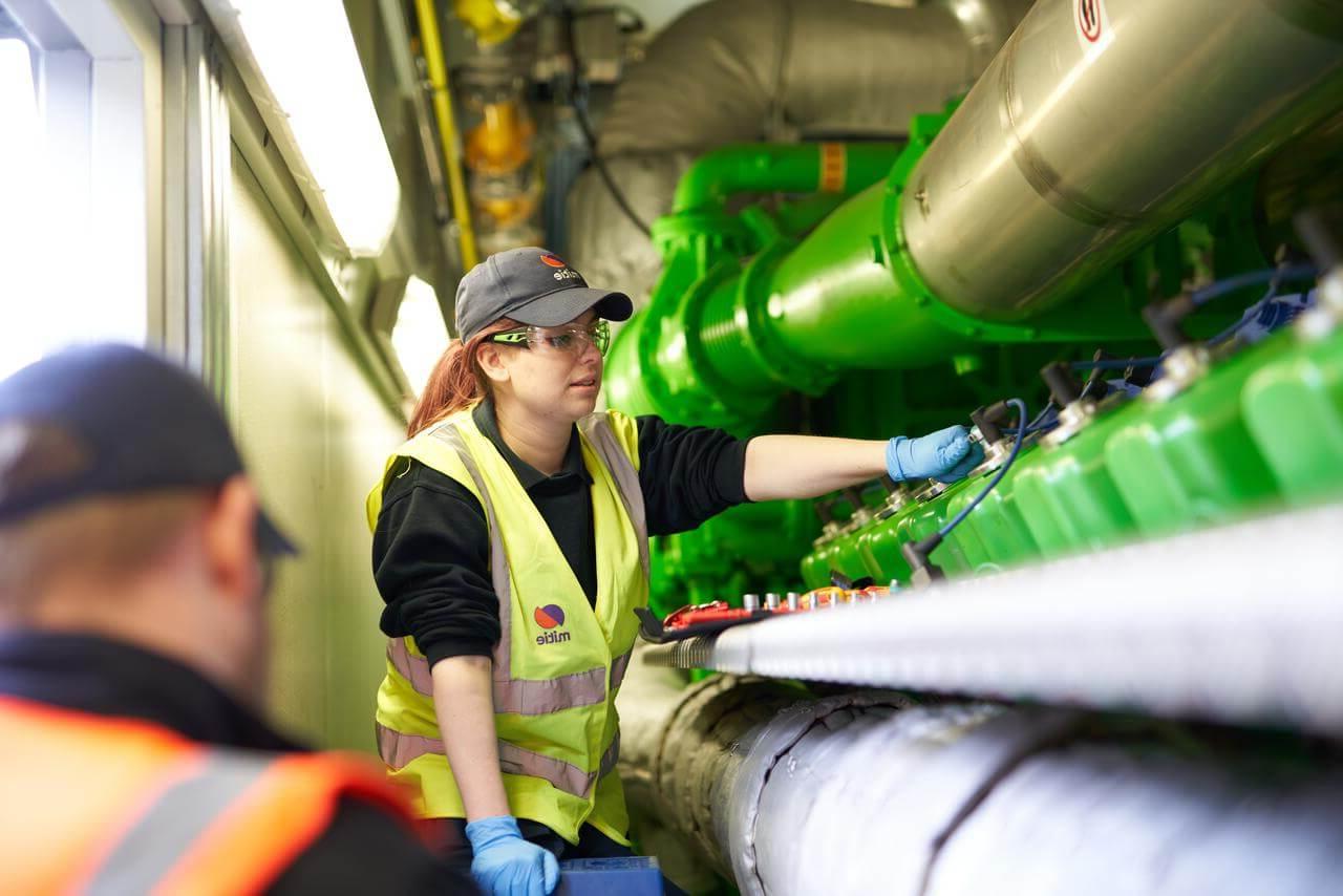 A female Mitie engineer wearing high-vis and safety glasses, working on pipes and equipment